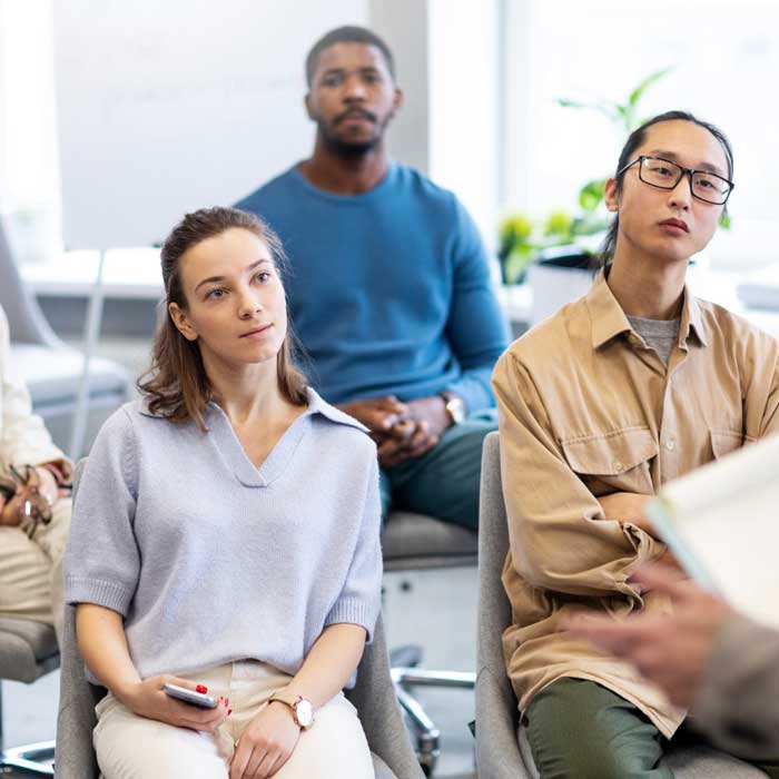 Life Coaching session showing a small group of adults attentively listening to a coach during a personal development workshop in a modern indoor setting.